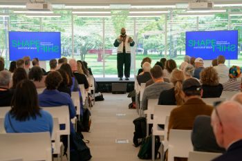 A man on a dais speaks with a microphone to dozens of people seated. Screens on both sides of him say "Shapeshift Global Summit Responsible AI Institute of Design." Behind the man is a wall of glass showing greenery and a sunny day outside.